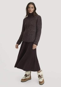 Dunkellila Strickpullover mit hohem Kragen, kombiniert mit einem fließenden dunkellila Rock. Das Model trägt braune Stiefel mit weißen Fellakzenten.