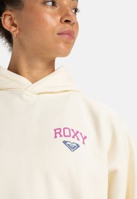 Krémová mikina z mäkkého materiálu s ružovým logom "ROXY" a modrým srdcom nižšie, navrhnutá v klasickom uvoľnenom strihu s kapucňou.