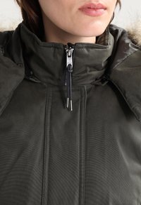 Veste d'hiver vert foncé avec un col montant, une fermeture éclair visible et un cordon de serrage. Dotée d'une texture matelassée et d'une bordure en fourrure au niveau du col.