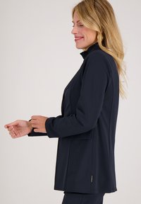 Marine softshell blazer met hoge kraag, voorzakken en een gladde textuur. Heeft een zijsplit detail en een subtiele merketiket aan de zoom.
