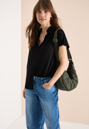 Ung kvinde iført sort kortærmet skjorte med knapper og blå jeans, bærende en quiltet olivengrøn skuldertaske med den ene hånd i lommen.