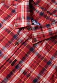 Camisa de franela a cuadros en rojo y azul con cuello abotonado. Presenta botones marrones y una etiqueta que dice "Superdry". Textura suave y material cálido.