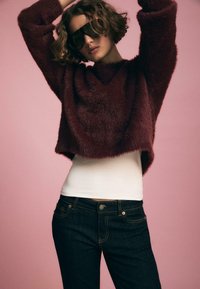 Bordeauxfarbener, flauschiger Pullover mit weitem Ausschnitt, kombiniert mit einem weißen Tanktop und dunkelblauen Jeans mit gelben Nähten und Fronttaschen.