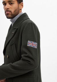 Donkergroene dubbelzijdige jas met grote revers, gouden knopen en een Union Jack-patch op de linker mouw. Glad textuur en getailleerde pasvorm.
