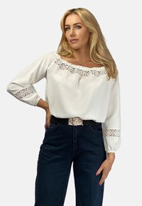 Biała bluzka z koronkowym wykończeniem przy dekolcie i mankietach, zestawiona z ciemnoniebieskimi dżinsami z wysokim stanem oraz czarnym paskiem z metalowymi elementami.