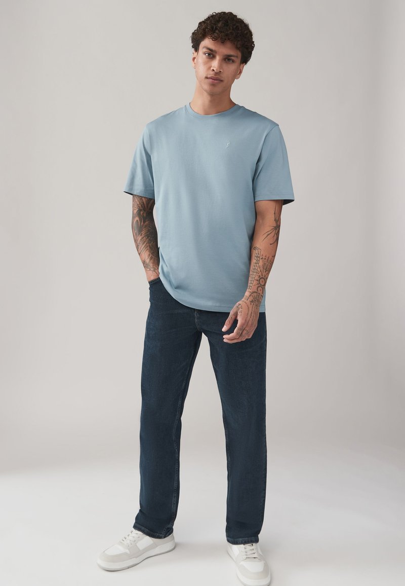 Giovane uomo con capelli ricci che indossa una maglietta azzurro chiaro, jeans scuri, scarpe da ginnastica bianche e tatuaggi sulle braccia, in piedi davanti a uno sfondo semplice.