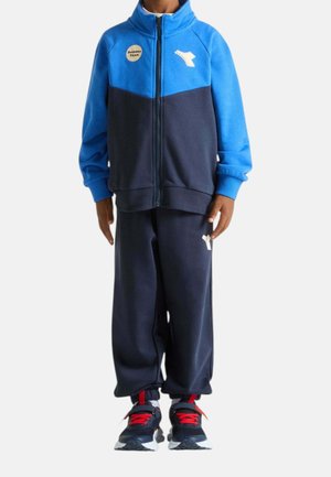Bambino con tuta blu e blu navy con zip frontale e scarpe da ginnastica blu navy con lacci rossi, in piedi davanti a uno sfondo semplice.