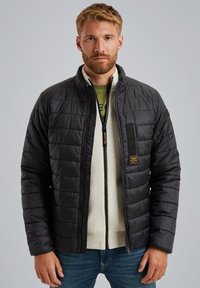 Chaqueta negra acolchada con cuello alto, cierre de cremallera y bolsillo en el pecho. Lleva puesta un suéter de color crema claro, con un patrón texturizado.