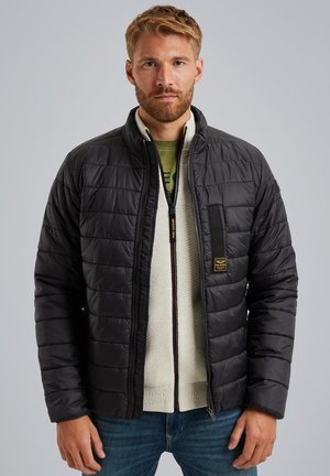 Giacca nera trapuntata con colletto alto, chiusura con zip e tasca sul petto. Indossata sopra un maglione color crema chiaro, con un motivo testurizzato.