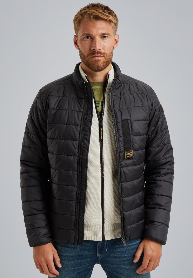 Chaqueta negra acolchada con cuello alto, cierre de cremallera y bolsillo en el pecho. Lleva puesta un suéter de color crema claro, con un patrón texturizado.