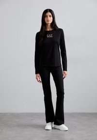 Zwarte blouse met lange mouwen en logo-detail, gecombineerd met zwarte uitlopende broek en witte sneakers. Glad stof, slimfit design, geen patronen.
