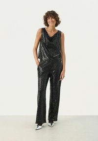 Sort jumpsuit med pailetter, draperet halsudskæring og vid buksestil. Har lommer og sølvfarvede spidse hæle. Glat, skinnende tekstur hele vejen igennem.