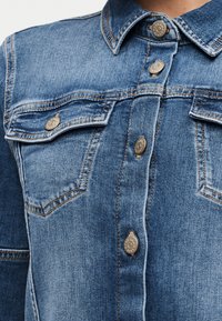 Κοντινή λήψη μπροστινού μέρους μπλε τζιν μπουφάν με μεταλλικά κούμπωμα κουμπιά και θήκες στο στήθος με καπάκι, ραμμένες με πορτοκαλί ραφές.