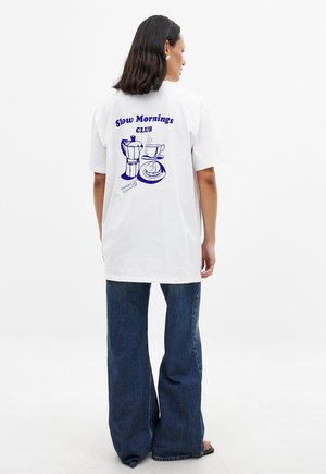 Weißes T-Shirt mit blauer Grafik eines Kaffeebereiters und einer Tasse auf der Rückseite. Lässiger Schnitt mit kurzen Ärmeln, kombiniert mit ausgestellten Jeans.