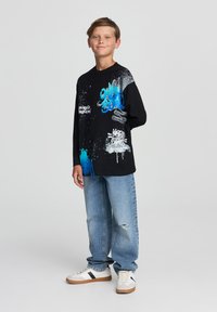 Maglietta nera a maniche lunghe con motivi di graffiti colorati, abbinata a jeans chiari strappati e sneakers beige con accenti neri.