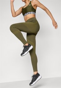Olívazöld sportmelltartó és hozzá illő leggings texturált részletekkel; fekete-fehér sportcipővel párosítva, a modell dinamikus pózban.