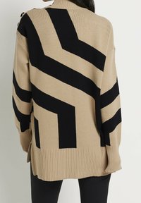Beige gebreide trui met opvallende zwarte zigzagpatronen, geribde zoom en knoopaccenten op de schouder. Glad materiaal, ontspannen pasvorm.