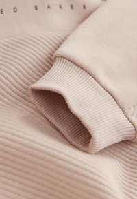 Šviesiai rožinės spalvos džemperio audinys su tekstūruotais siaurais akcentais rankogalyje ir lygiu pagrindu. Ant audinio tamsiomis raidėmis išraižytas prekės ženklas.