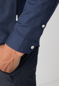 Personne portant une chemise bleu marine à manches longues avec poignets boutonnés, main reposant dans la poche d’un pantalon foncé sur un fond clair uni.