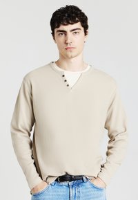Camicia beige a maniche lunghe con tessuto testurizzato, design a V, tre bottoni marroni come dettagli, indossata sopra una maglietta bianca, abbinata a jeans blu.