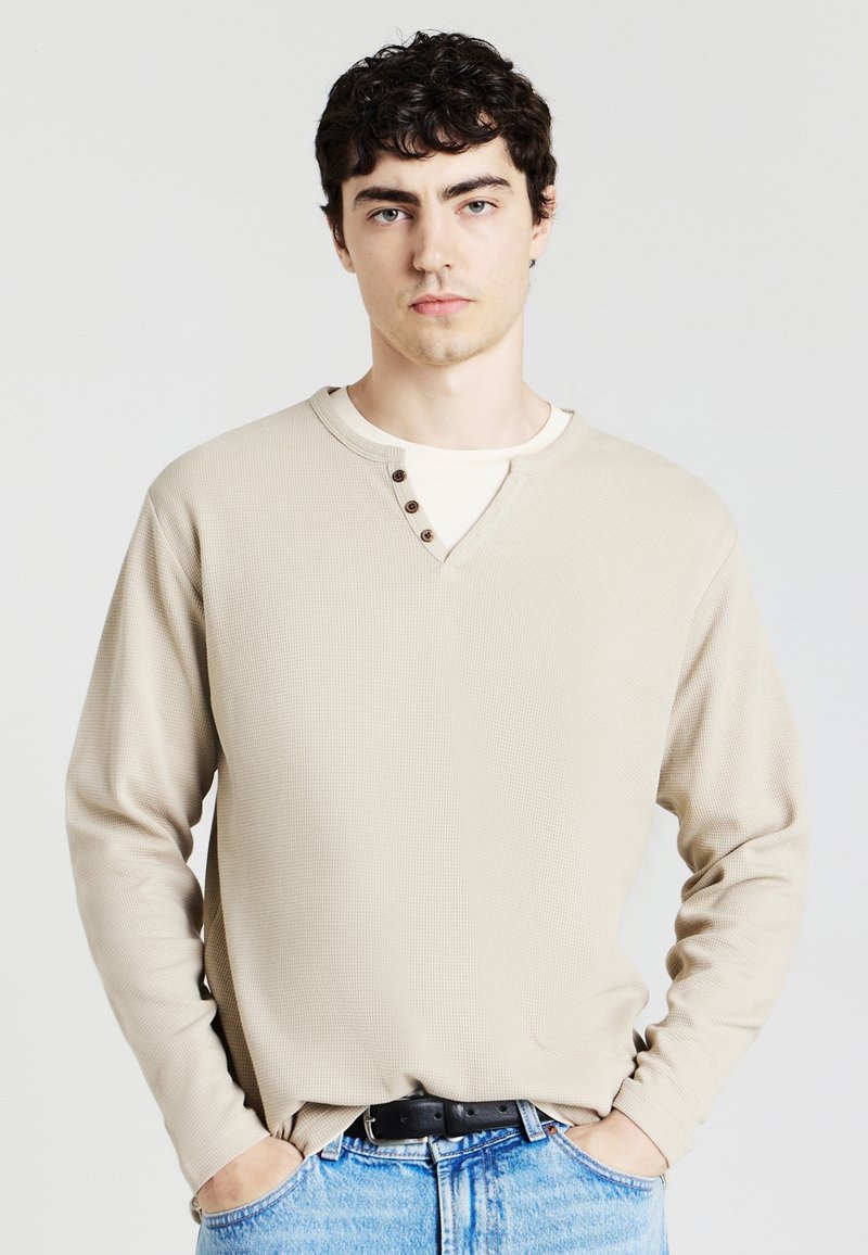 Camicia beige a maniche lunghe con tessuto testurizzato, design a V, tre bottoni marroni come dettagli, indossata sopra una maglietta bianca, abbinata a jeans blu.