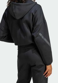 Svart huvtröja med vida ärmar, prydd med en reflekterande rand på vänster arm. Tillverkad av slät, matt tyg.