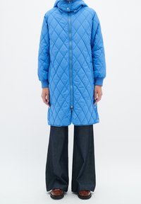 Abrigo acolchado azul con capucha, cierre frontal de cremallera, puños de canalé y botones de presión en los laterales. Llevado sobre unos pantalones oscuros de pierna ancha y zapatos marrones.