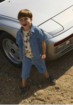 Denim oblačilo s kratkimi rokavi in cargo hlačami svetlo modre barve, dopolnjeno s kremasto majico z odtisi vozil in črnimi sandalni.