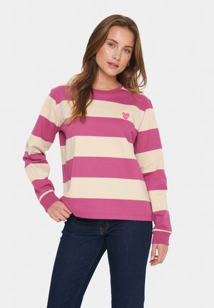 Donna con una maglietta a maniche lunghe a righe larghe rosa e beige con un piccolo emblema a forma di cuore, abbinata a jeans blu scuri, in posa su uno sfondo bianco.