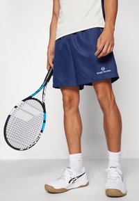 Navy athletic shorts with a logo, white shoes with black accents, white socks, and a tennis racket with a blue detail. Smooth fabric texture.