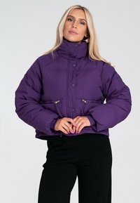 Fioletowa kurtka puchowa z wysokim kołnierzem, zamykanymi na zamek kieszeniami z przodu i elastycznymi mankietami. Charakteryzuje się skróconym fasonem oraz gładką, błyszczącą teksturą.