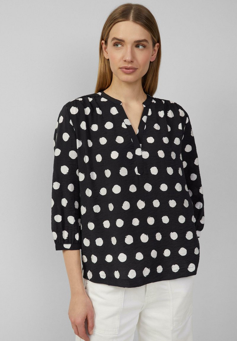 Zwarte shirt met witte stippen, lichte stof, 3/4 mouwen, V-hals en plooien op de schouders. Het patroon heeft een handgeschilderd effect.