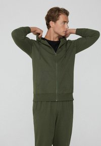 Sudadera con cremallera en verde oliva, fabricada en un tejido suave, que cuenta con un bolsillo frontal, cordones ajustables y un corte relajado, combinada con pantalones a juego.
