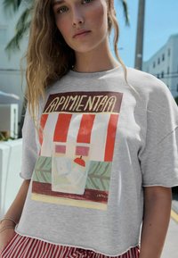 T-shirt grigia a maniche corte con una stampa grafica colorata di un negozio, con strisce rosse e foglie verdi nella parte inferiore.
