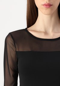 Juodas ilgos rankovės marškinėlis su permatomu tinklo iškirpimu, turintis apvalų apykaklę ir lygų, priglustantį siluetą. Minkšta tekstūra, nėra matomų raštų ar detalių.