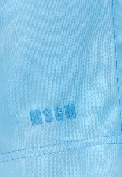 Κοντινό πλάνο ανοιχτό μπλε υφάσματος με ραμμένα γράμματα "MSGM" σε νήμα ίδιου χρώματος κοντά σε ραφή.