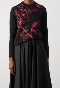 Black sweater with red and gold floral embroidery, round neck, and long sleeves. Worn over a textured black skirt.