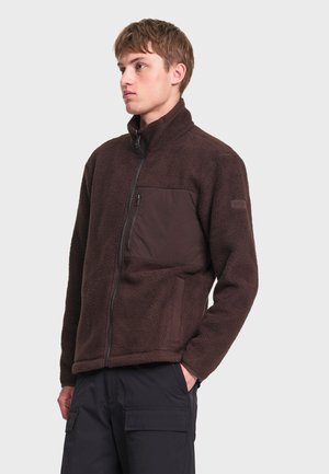 Jeune homme portant une veste polaire marron foncé avec une poche poitrine zippée et un pantalon cargo noir, debout, une main dans la poche.