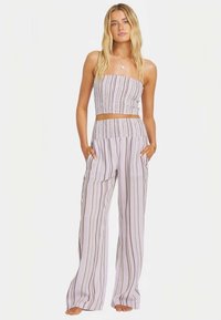 Ung kvinde står barfodet iført et lavendel to-deles outfit med lodrette brune striber og hænderne i lommerne.