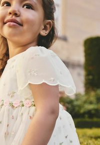 Weißes Kleid aus transparentem Stoff, mit bestickten pastellfarbenen Blumen, einem wellenförmigen Kappenärmel und einem rosa Blumenbesatz in der Taille.