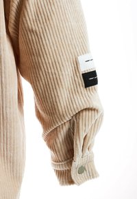 Manga de chaqueta de pana beige con textura acanalada, que presenta etiquetas de tela blanca y negra, y un botón a presión plateado en el puño.