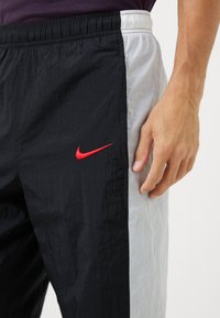 Nike træningsbukser med en sort og lysegrå farveskala. Har en elastisk talje, et rødt broderet logo og en let, krøllet tekstur.