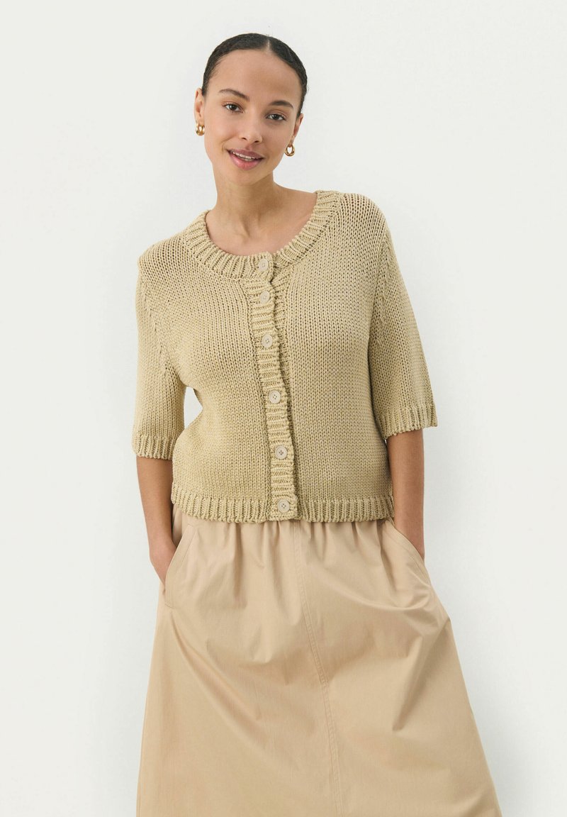Cardigan beige en tricot à manches courtes, col rond et fermeture à cinq boutons. Associé à une jupe beige fluide, dotée de poches latérales.