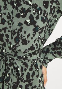 Grön klänning med leopardmönster, som har svarta, teal och vita fläckar. Tyget är mjukt med detaljer i knytmidja och knappstängning fram.