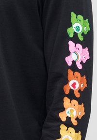 Manche noire avec cinq Care Bears colorés en vert, rose, marron, orange et jaune, chacun avec un badge de ventre unique en rangée verticale.