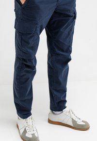 Marine cargobroeken gemaakt van lichtgewicht stof met meerdere zakken en een relaxte pasvorm, gecombineerd met witgrijze sportschoenen.