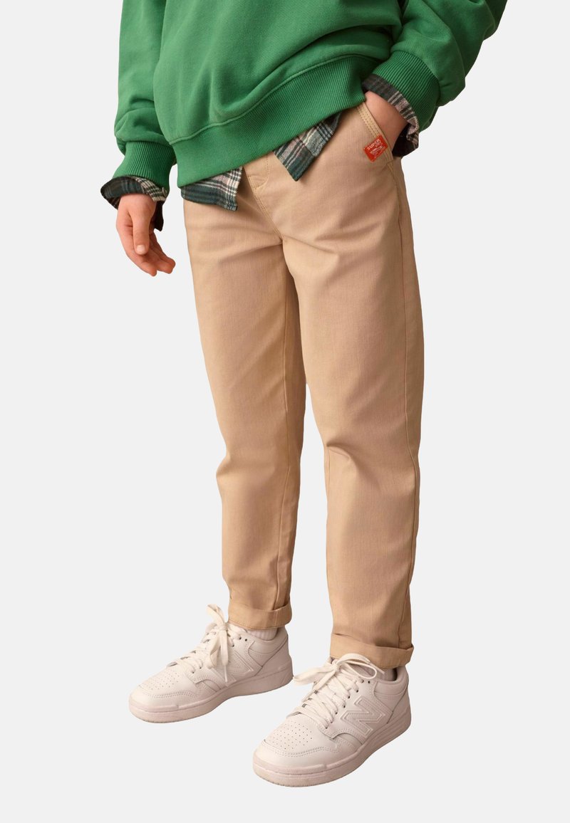 Chinos beiges à coupe droite, avec des coutures discrètes, des poches latérales et un ourlet roulé, associés à des baskets blanches et un sweat-shirt vert.