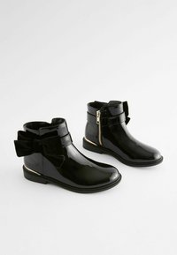 Schwarze Lackleder-Stiefeletten mit runder Zehenform, seitlichem Reißverschluss und einer großen Stoffschleife auf der Rückseite. Detail in goldfarbener Hardware.
