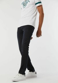 Jeans foncés ajustés associés à un t-shirt blanc avec un graphique vert et des manches rayées, portés avec des baskets blanches.