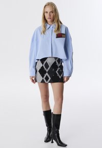 Camicetta azzurro chiaro a maniche corte con taschino a quadretti, abbinata a una minigonna nera e bianca con motivo a rombi e spille da balia. Stivali in pelle nera.